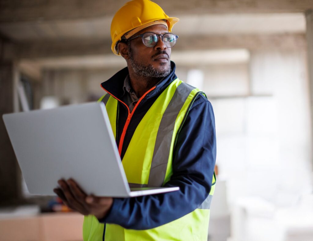A man standing in high vis wearing a yellow safety hat using a laptop to manage his business